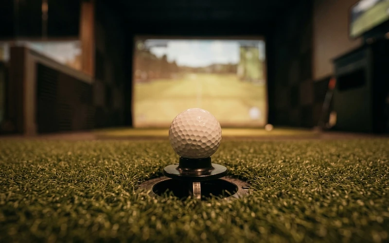 Close-up of a golf ball resting on an automatic tee mechanism set into artificial turf in a golf simulator bay.