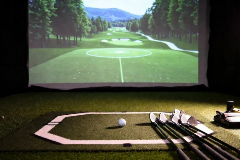 Home golf simulator setup with a row of irons and a ball on a hitting mat, a launch monitor, and a fairway projected on the impact screen.