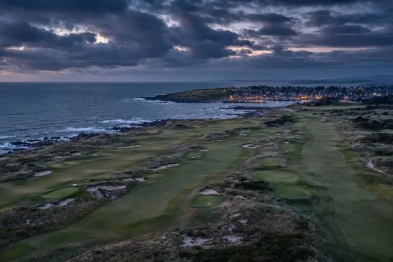 Aerial view of a coastal links golf course at dusk with a harbour town lit in the distance